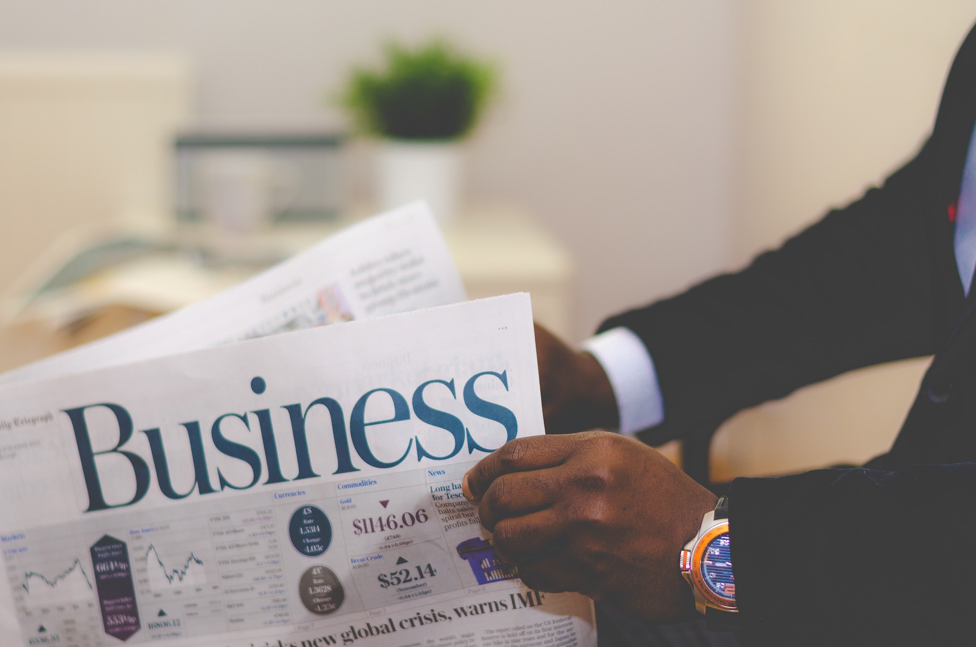 Person reading a business newspaper with financial charts and headlines visible. A potted plant sits on a table in the background.