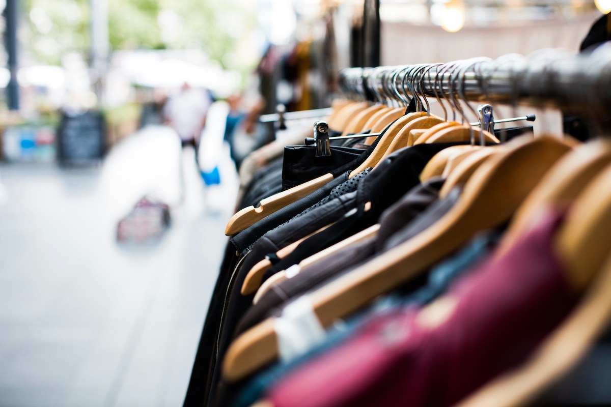 A row of assorted clothes on wooden hangers hangs on a metal rack in an outdoor market setting.