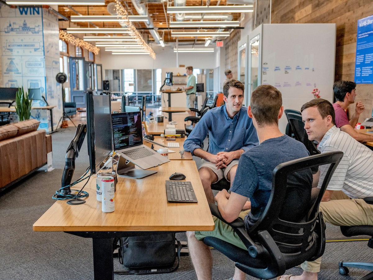 A group of people work and converse at a long desk in a modern, open-plan office with wooden accents and ample natural light. Computers, monitors, and office supplies are visible on the desk.