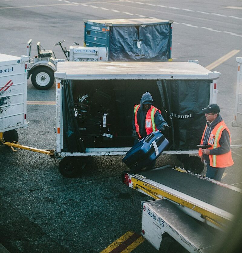 Two airport baggage handlers dressed in safety vests load luggage into trailers on the tarmac.