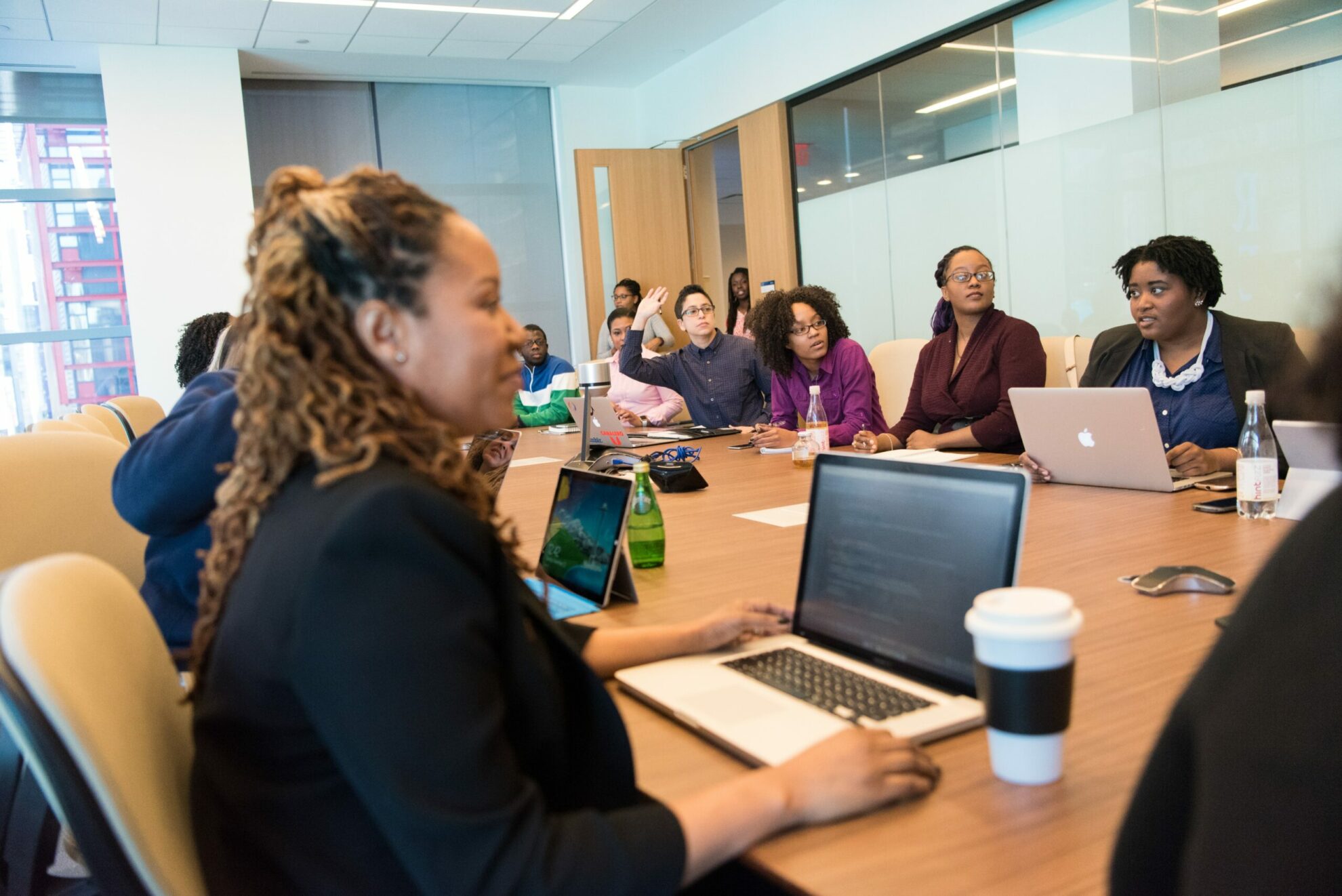 A group of people are seated around a large conference table in an office setting, some with laptops open, actively engaging in a meeting.