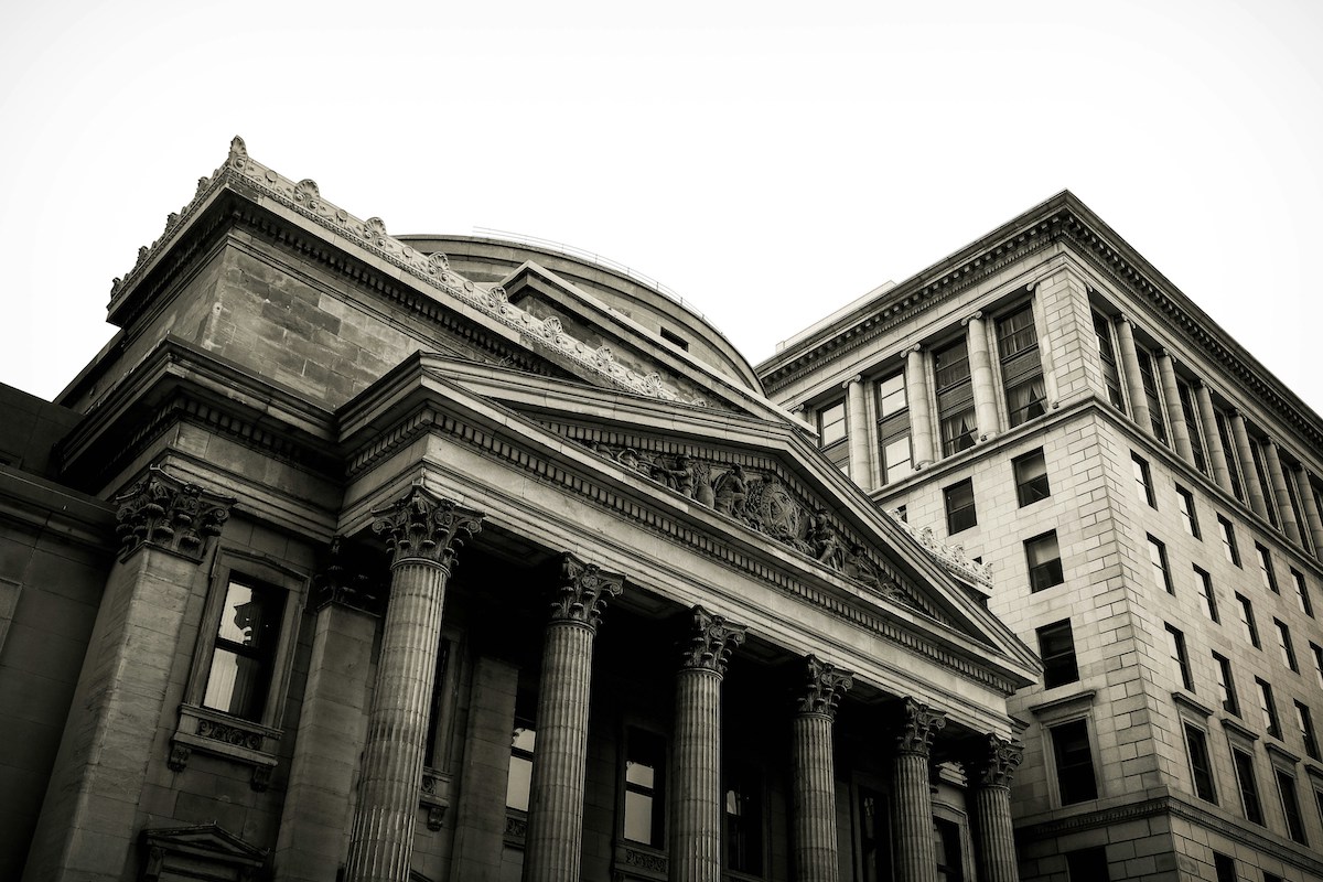 A black-and-white photo of a neoclassical building featuring tall columns and ornate architectural details next to a tall, rectangular, modern-looking building.