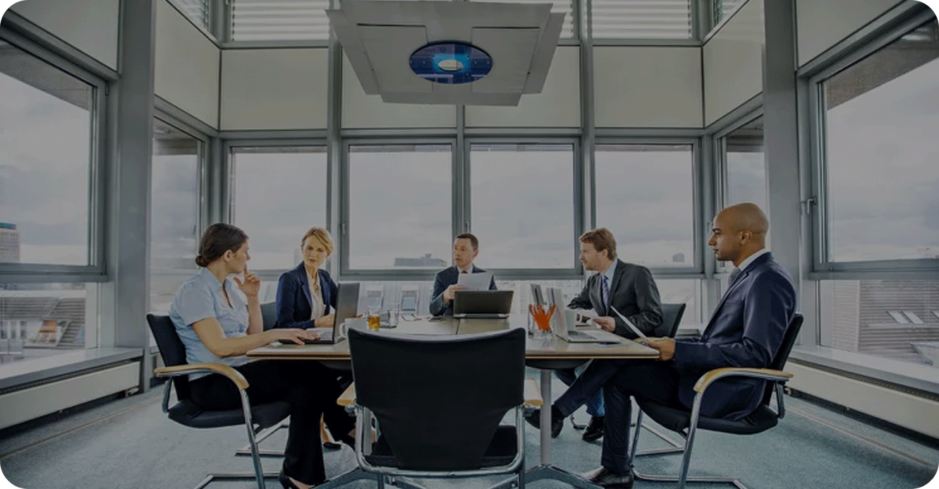 Five people in business attire sit around a conference table in a modern office with large windows, engaged in a discussion.