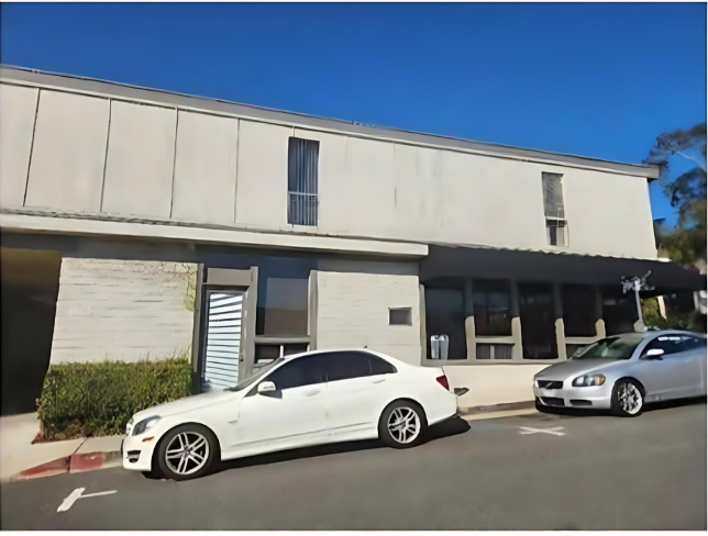 Two white cars are parked in front of a two-story building with a flat roof and vertical windows on a sunny day. The building has a light gray exterior and surrounding greenery.