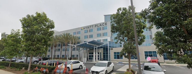 A multi-story Hyatt Place hotel with a glass entrance canopy, surrounded by trees and parked cars in the front lot.