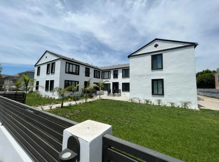 A modern, white two-story house with black-trimmed windows and a manicured lawn is surrounded by a black fence. The sky is partly cloudy.