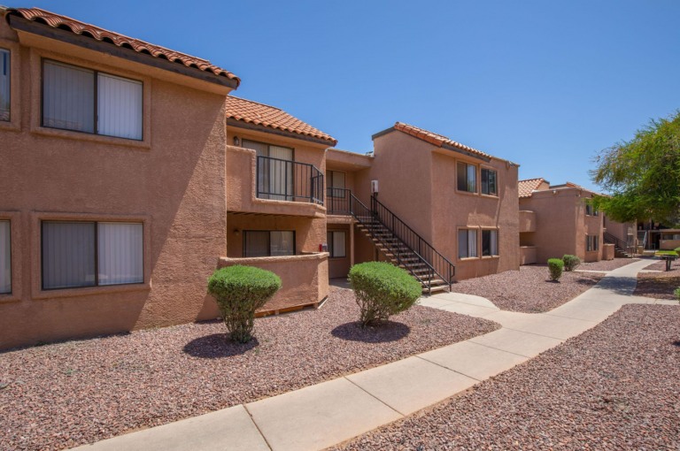 A beige stucco apartment building with red-tiled roofs, a central staircase, and well-maintained desert landscaping on a clear, sunny day.