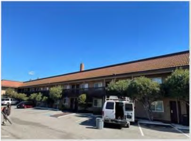 A two-story apartment building with a brown roof, viewed from a parking lot. There are several parked cars, including a white van, and some trees in front of the building. The sky is clear and blue.