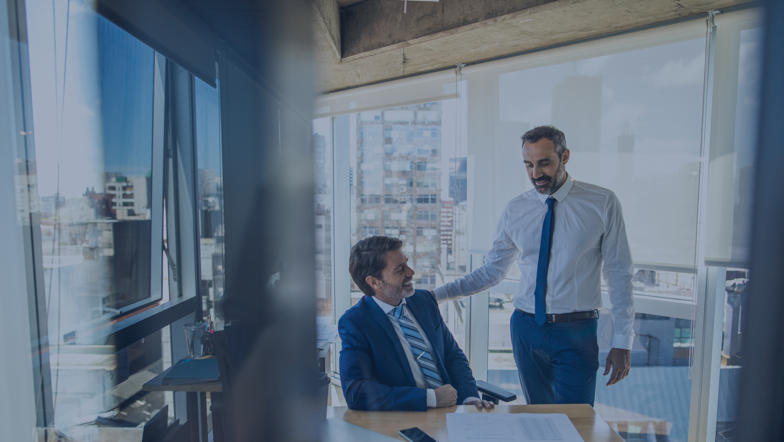 Two men in business attire are in an office. One is seated at a desk with papers; the other stands beside him with a hand on his shoulder. Large windows show a cityscape background.