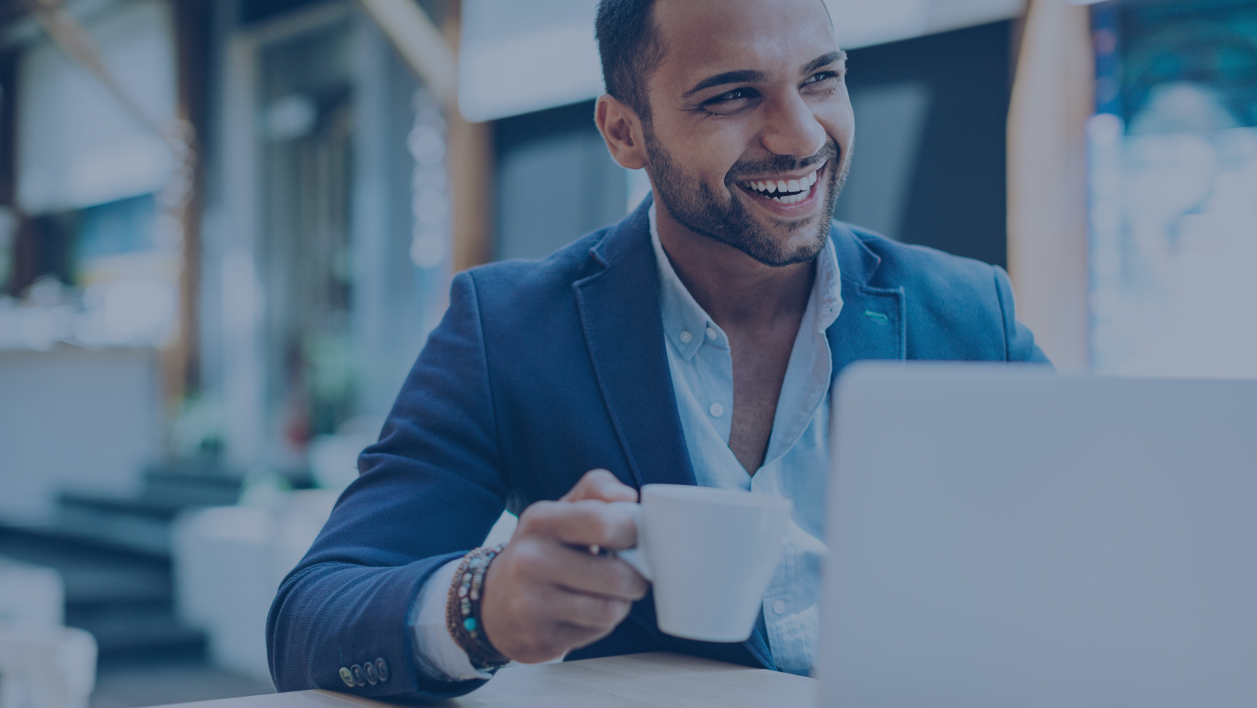 A person in a blue blazer is smiling while holding a cup and sitting at a desk with a laptop.