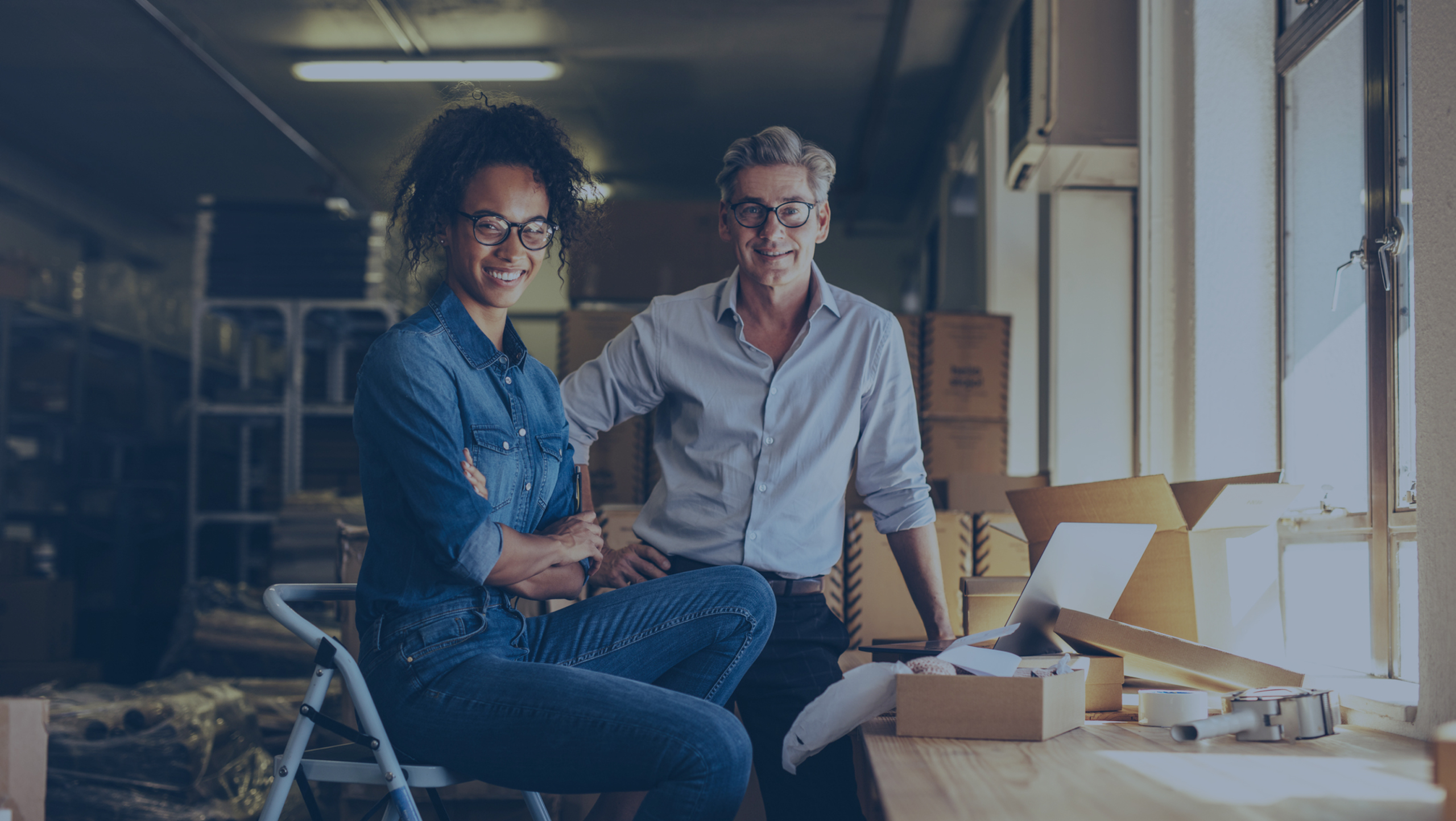 Two people smiling in a workspace with boxes and a laptop. One sits on a stool, while the other leans against the table.