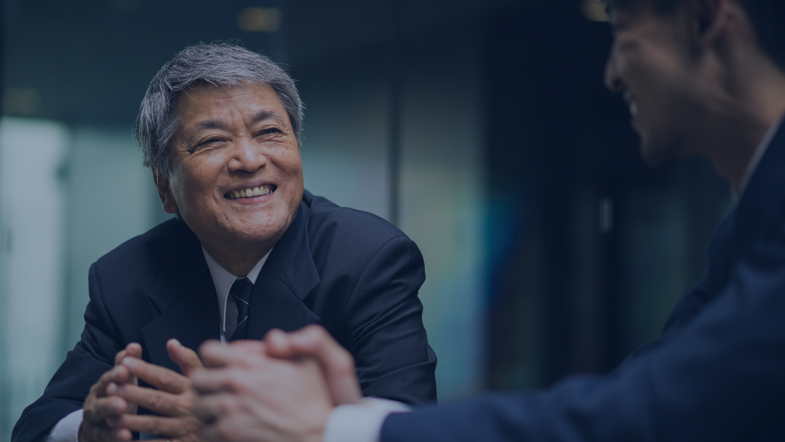 Two men in suits are having a conversation. The older man is smiling and clasping his hands.