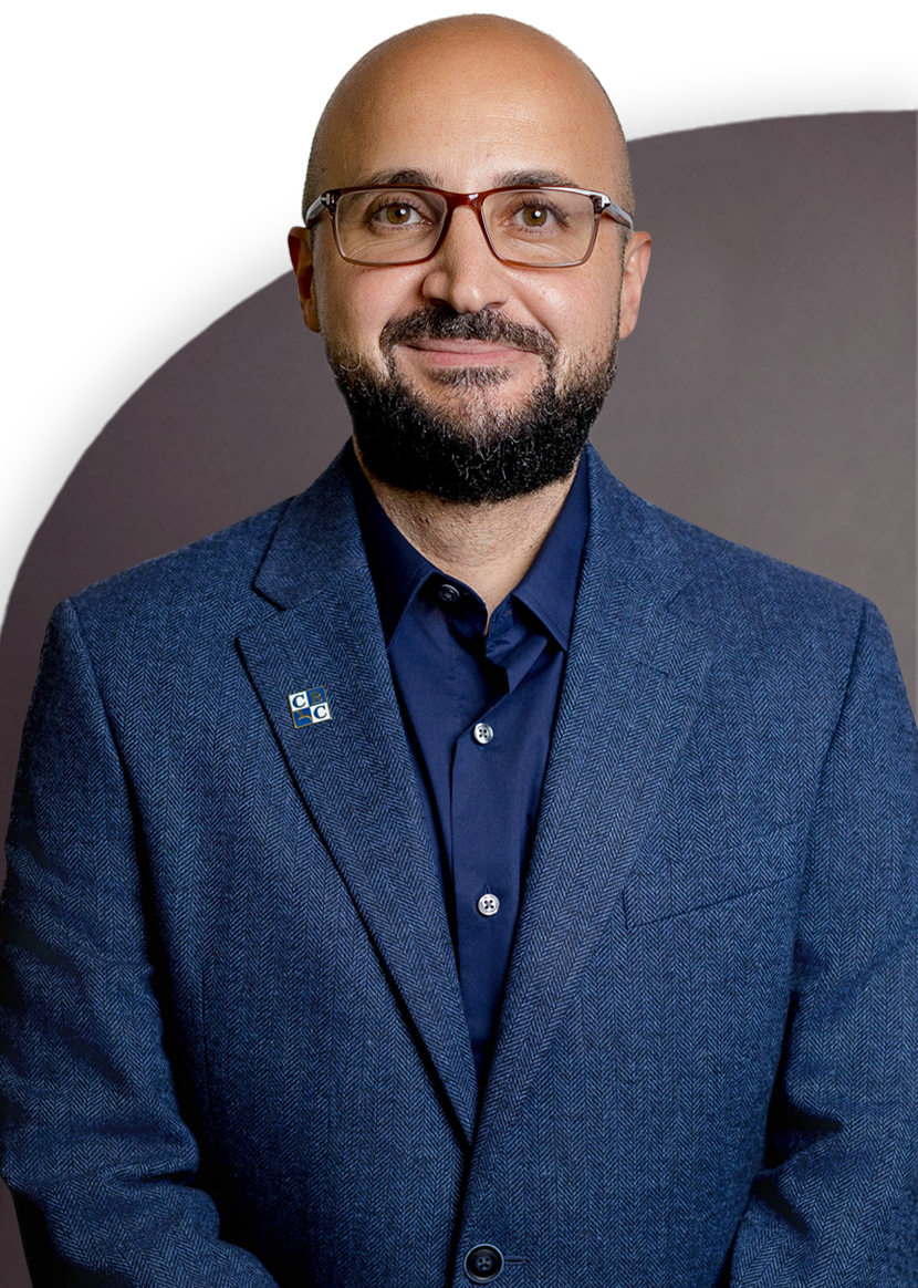 A person with a beard and glasses wearing a blue suit and a blue shirt, standing against a plain background.