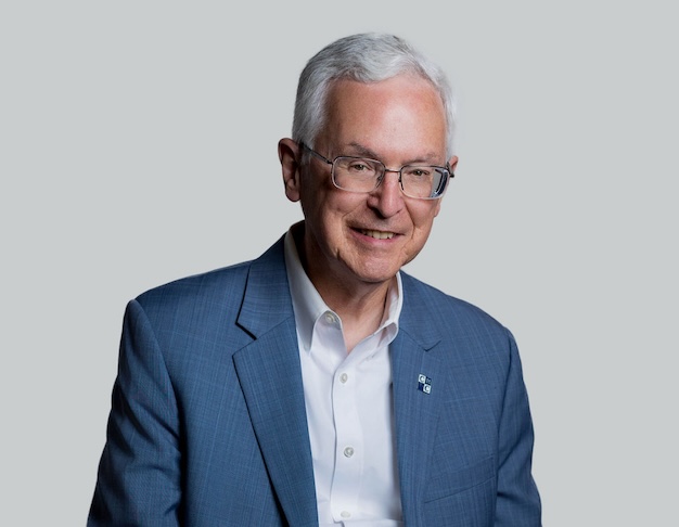 Older man with gray hair and eyeglasses, wearing a blue blazer and white shirt, smiles in front of a plain background.