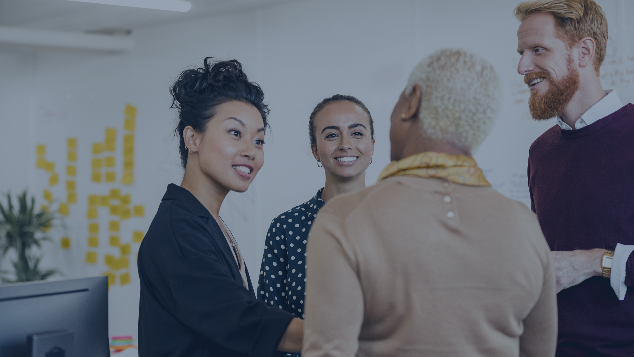 Four people stand in an office, engaged in conversation. Sticky notes are visible on the wall in the background.