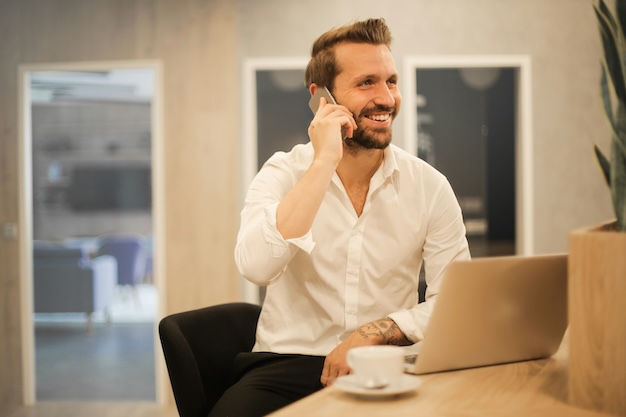 A man in a white shirt is smiling while talking on a smartphone, seated at a desk with a laptop and coffee cup.