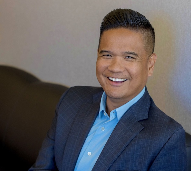 A person in a blue shirt and dark blazer sits on a couch, smiling at the camera.