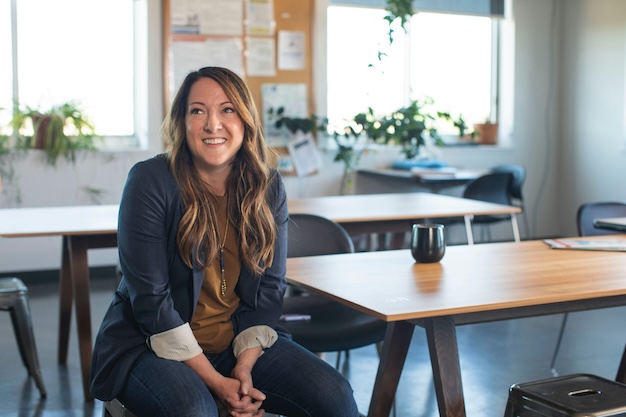 A woman sitting on a chair in an office with wooden tables and a mug. She is smiling, wearing a blazer, and there are plants in the background.