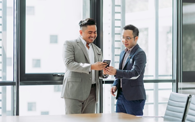 Two men in suits stand by a window, smiling while looking at a smartphone. A conference table and chairs are in the foreground.