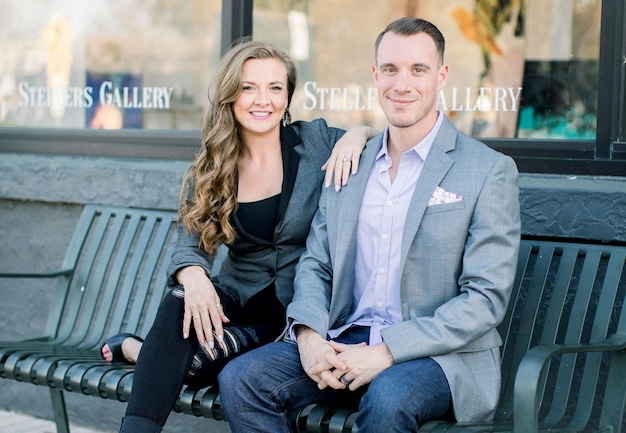 Two people sitting on a bench outside a gallery, both wearing casual business attire. The woman has long hair and is smiling, and the man is seated with his hands clasped.