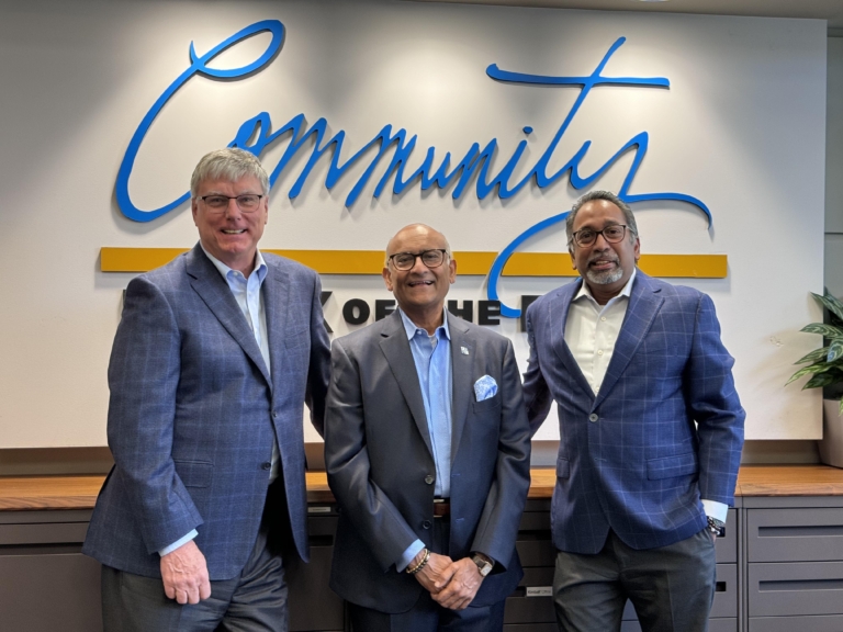 Three men in suits stand in front of a wall with the word "Community" written in large script. They are smiling and standing close together. A plant is visible on the right.