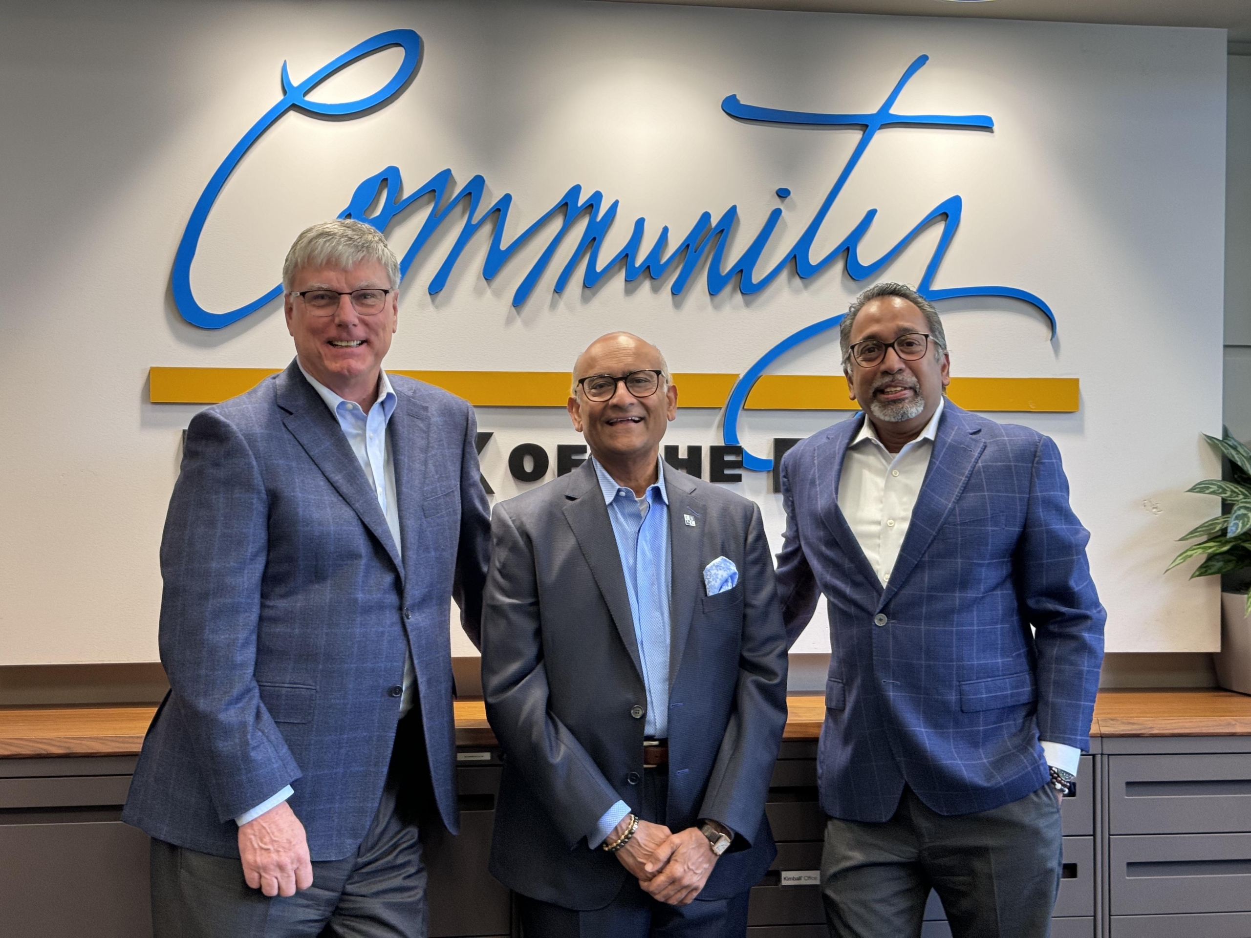 Three men in suits stand in front of a wall with the word "Community" written in large script. They are smiling and standing close together. A plant is visible on the right.