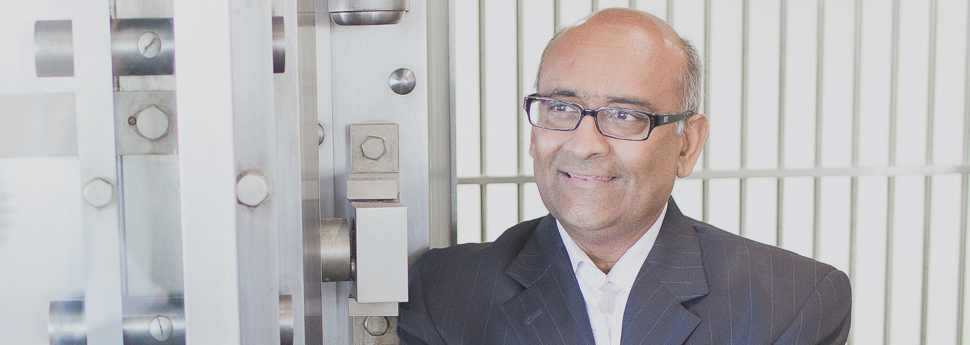 A man in a suit and glasses stands beside a large metal structure with bolts.