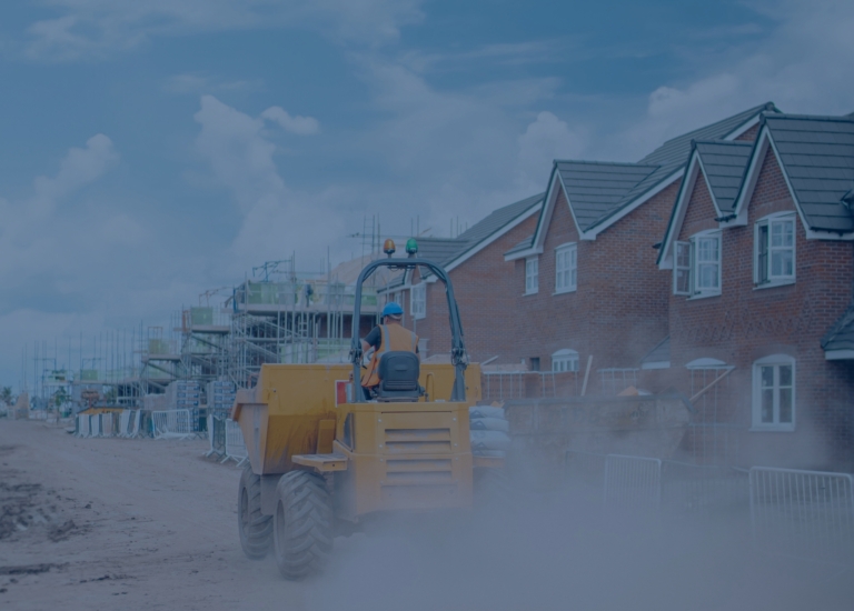 A construction worker operates a large yellow vehicle on a dusty road near partially built brick houses under a blue sky. Scaffolding is visible on the unfinished structures.