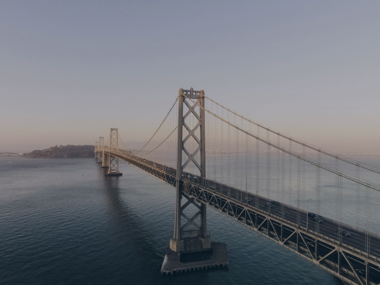 A large suspension bridge spans across calm water under a clear sky, with cars visible on the roadway and land in the distance.