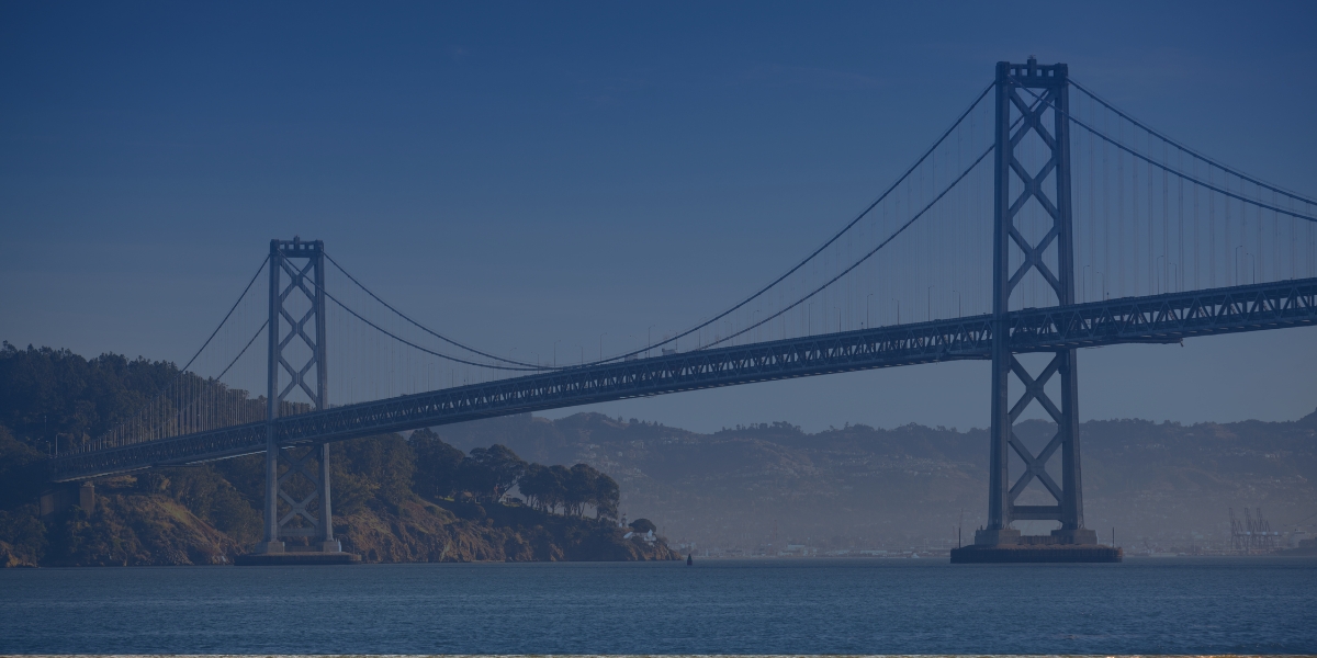 A suspension bridge spans across a body of water with hills in the background. The sky is clear and blue.