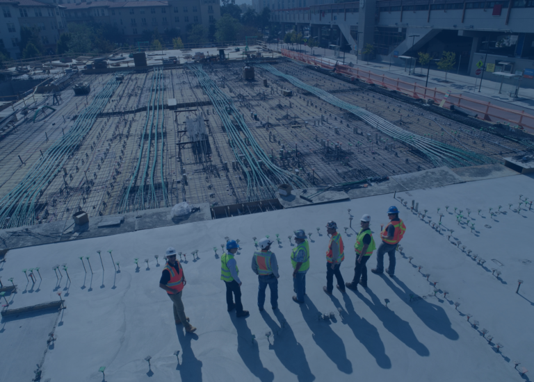 A group of construction workers wearing safety vests and helmets stand on a building site, surveying the housing starts progress with visible rebar and foundations.