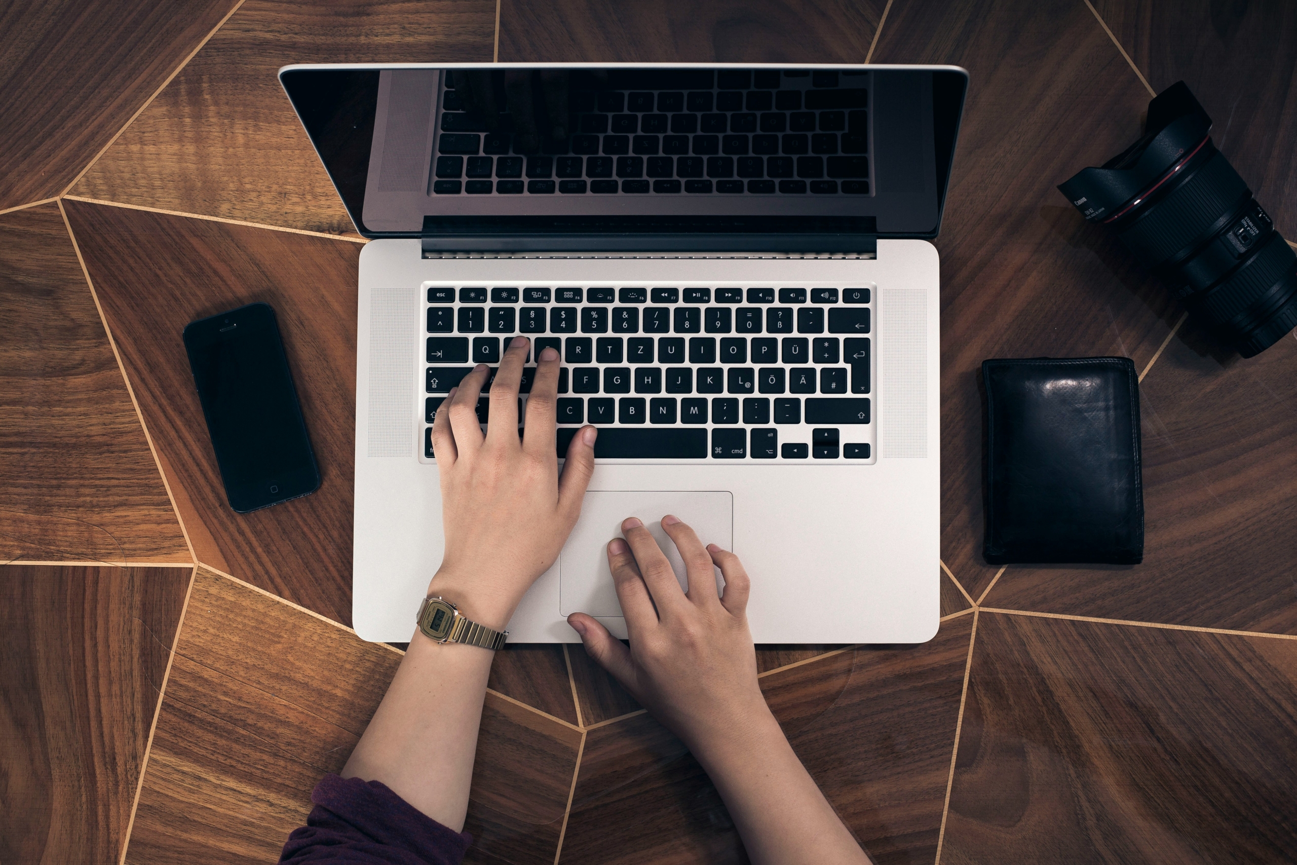 Hands typing on a laptop keyboard placed on a wooden surface, with a smartphone, camera lens, and wallet nearby.