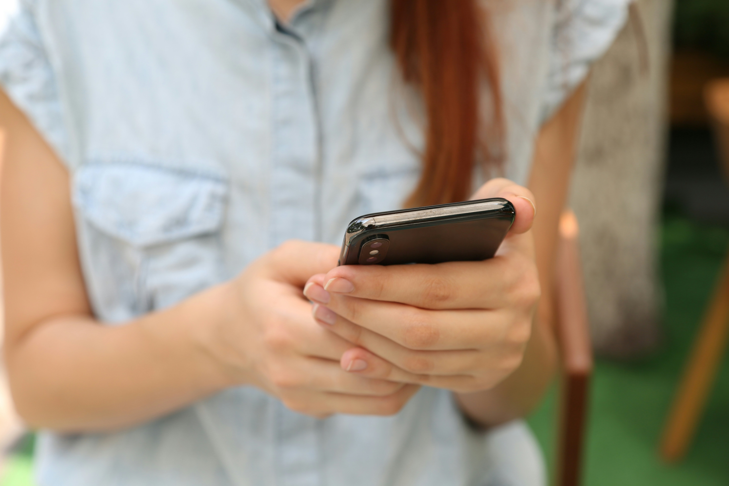 A person in a denim shirt holding and using a smartphone with both hands.