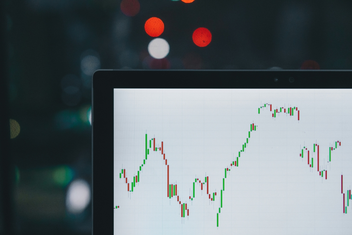 A computer screen displaying a stock market chart with red and green candlestick patterns in the foreground, home sales data subtly interwoven, and blurred lights in the background.