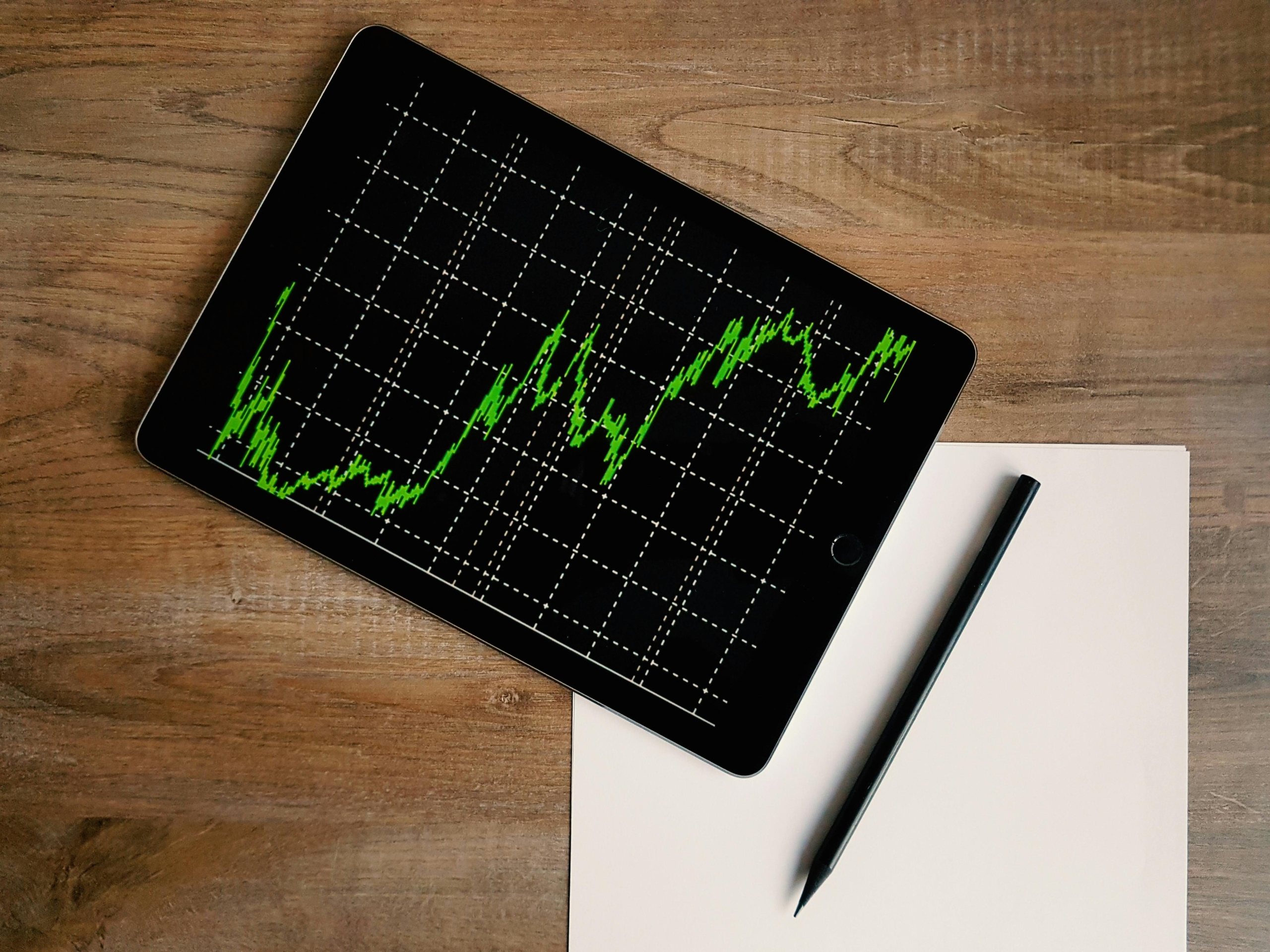 A tablet displaying a green line graph tracking interest rates lies on a wooden table next to a blank sheet of paper and a black pen.