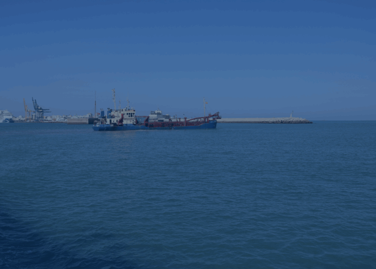 A large cargo ship is anchored in a calm harbor, with port cranes and a breakwater visible in the background under a clear blue sky, as shifting interest rates influence global shipping activity.